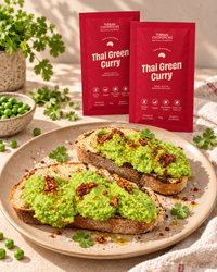 Two red packets of Urban Chefs Selects Thai Green Curry standing behind plated toast topped with green curry, garnished with herbs and red chili flakes, with bowls of peas and cilantro on a light fabric background.