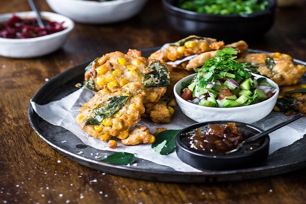A plate of golden-brown fritters with corn and green herbs, served with a side salad and dipping sauce on a dark plate on a wooden table.